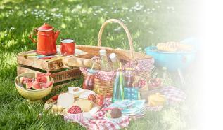 Image of a picnic hamper with food and drink on a blanket on the grass